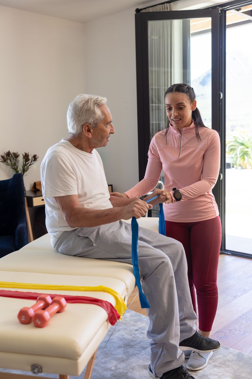 Mobile Physiotherapie als Hausbesuch in München – Physiotherapie Köpple Physiotherapeutin hilft einem älteren Patienten beim Üben mit dem Widerstandsband zuhause