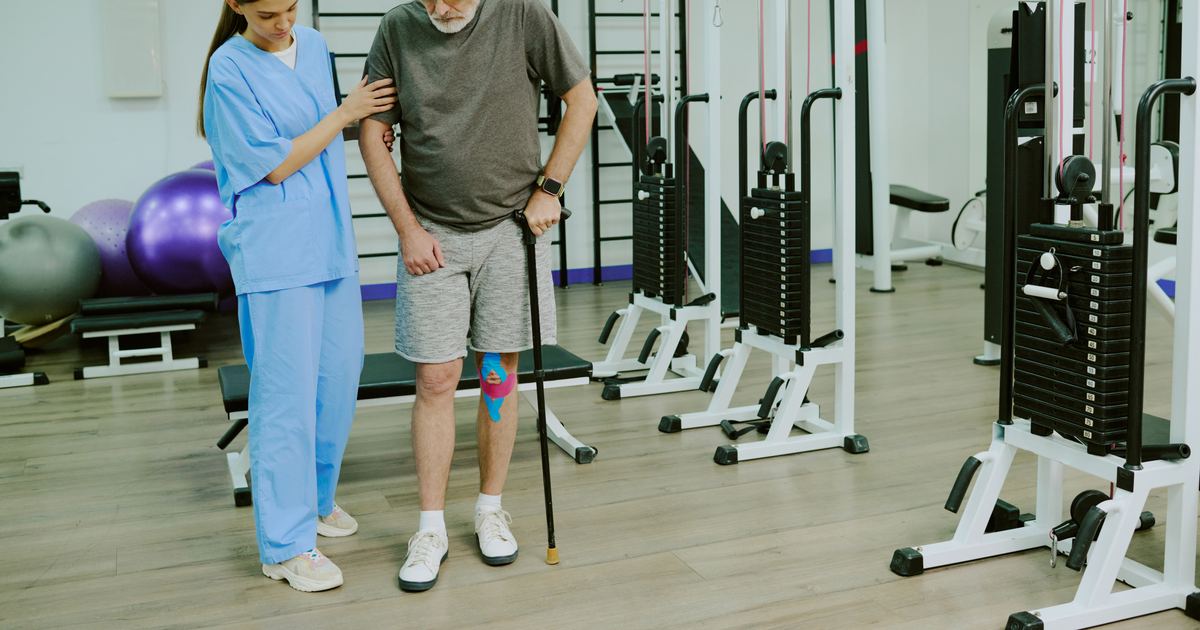 Physiotherapeutin unterstützt einen älteren Patienten Schritt für Schritt beim Gehtraining