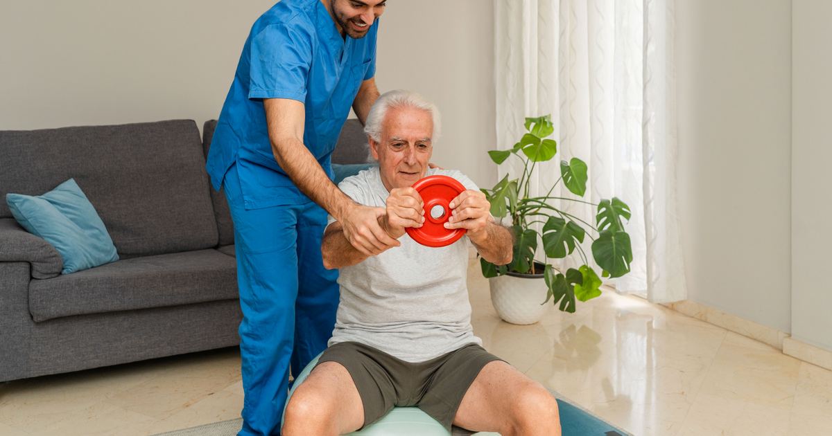 Krankengymnastik (KG) als Hausbesuch in München – Physiotherapie Köpple Senior trainiert unter Anleitung eines Physiotherapeuten mit einem Übungsgerät in seinem Wohnzimmer